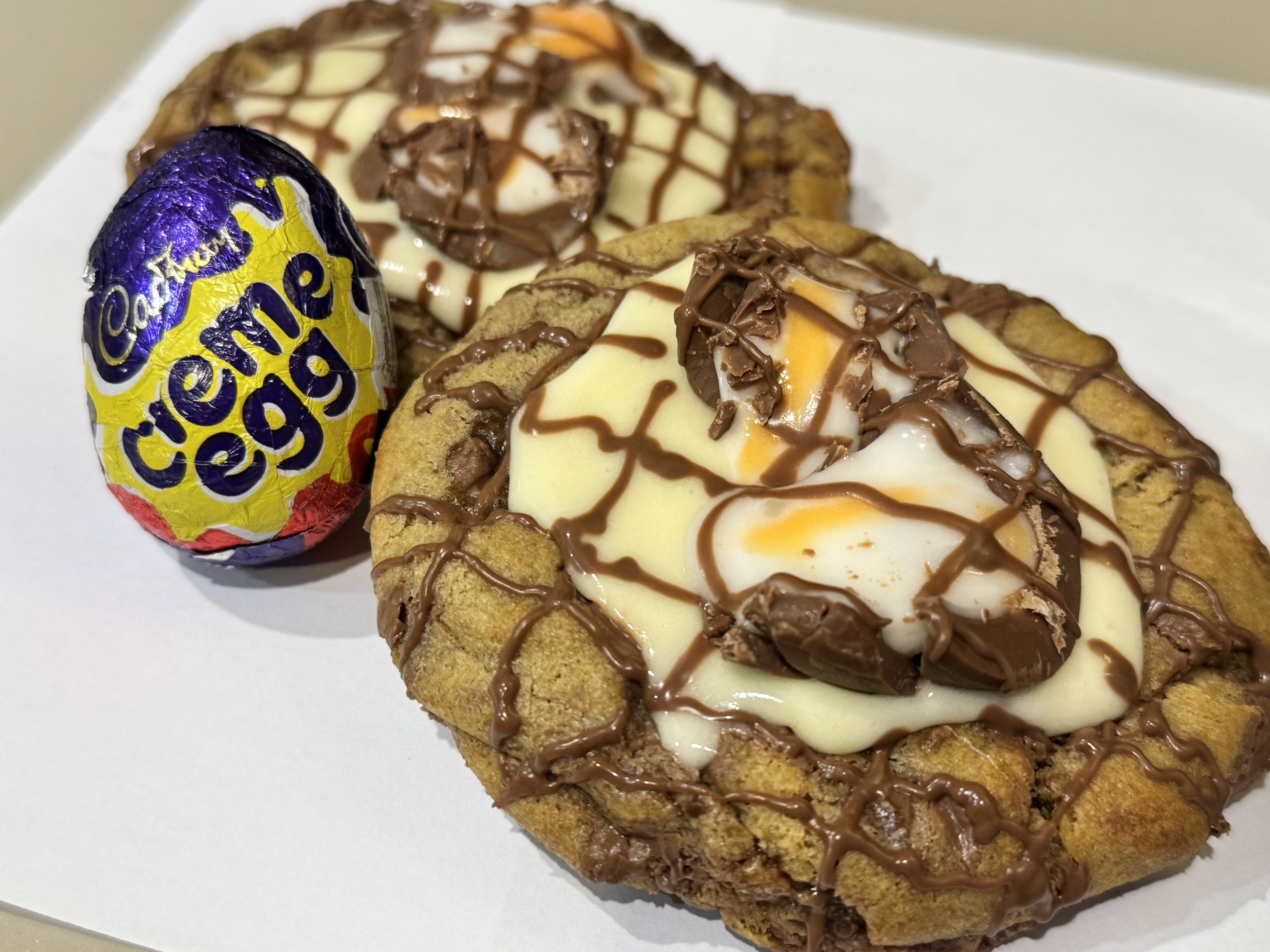 Two thick cookies are topped with melted white‑and‑orange fondant, drizzled milk chocolate, and half a Cadbury Creme Egg. A whole foil‑wrapped Creme Egg sits beside them.